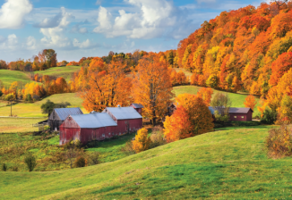 A group of red farm buildings located on rolling hills bordered by trees with yellow and orange leaves. 