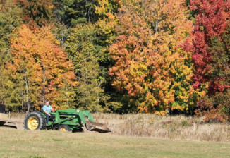 Parents and children ride in a trailer pulled by a tractor in a large field surrounded by trees in fall.