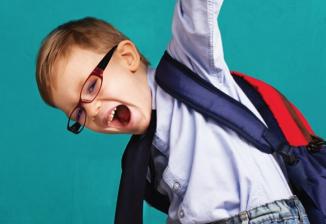 A young boy wearing glasses and a backpack smiles while raising one arm in the air.