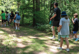Campers with white canes walk down a tree lined forest path. 