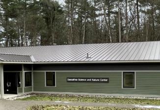 The Sassafras Science and Nature Center in a clearing in the woods. The rectangular building is painted sage green on the bottom and dark brown at the top.