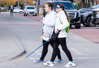 A girl with a white cane and a friend walk across a city street. 