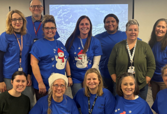 A group of fourteen teacher consultants pose for a group photo in a large room, mostly wearing matching blue shirts.
