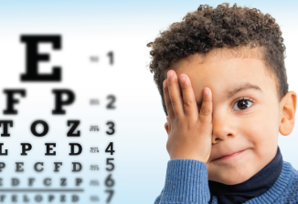 Young child in a blue sweater covering one eye, next to an eye chart during an eye exam.