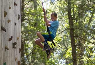 A boy smiles while rappelling down a wooden climbing wall in the woods.