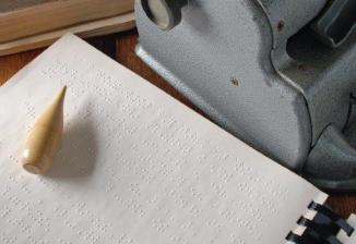 A braille writer rests on a table next to a sheet of braille and other braille tools.