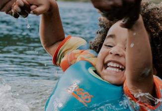 A young child laughs while playing in the water and holding onto an adult's hands.