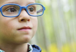 A young boy wearing glasses and a hearing device smiles while standing outside.