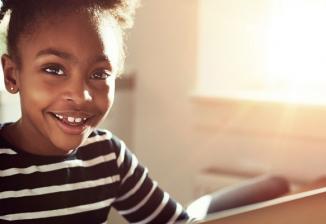 A young girl smiles at the camera while sitting in front of a laptop at home.