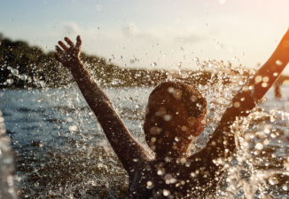 A young boy joyfully raises his hands in the air while swimming in a lake.