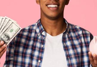 A smiling teenager holds a handful of paper money in one hand and a piggy bank in the other.