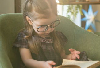 A young girl wearing glasses looks down at a book she is reading while sitting in a chair.